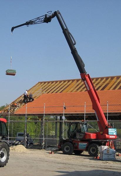 Ein Roter Manitou Teleskopkran, der ein großes Objekt anhebt, vor einer ländlichen Kulisse.