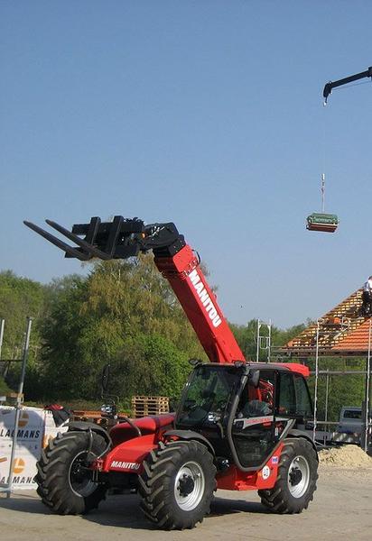 Ein Roter Manitou Teleskoplader, der ein großes Objekt anhebt, vor einer ländlichen Kulisse.